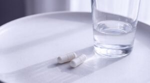 Two plain white capsules beside a glass of water on a matte white surface